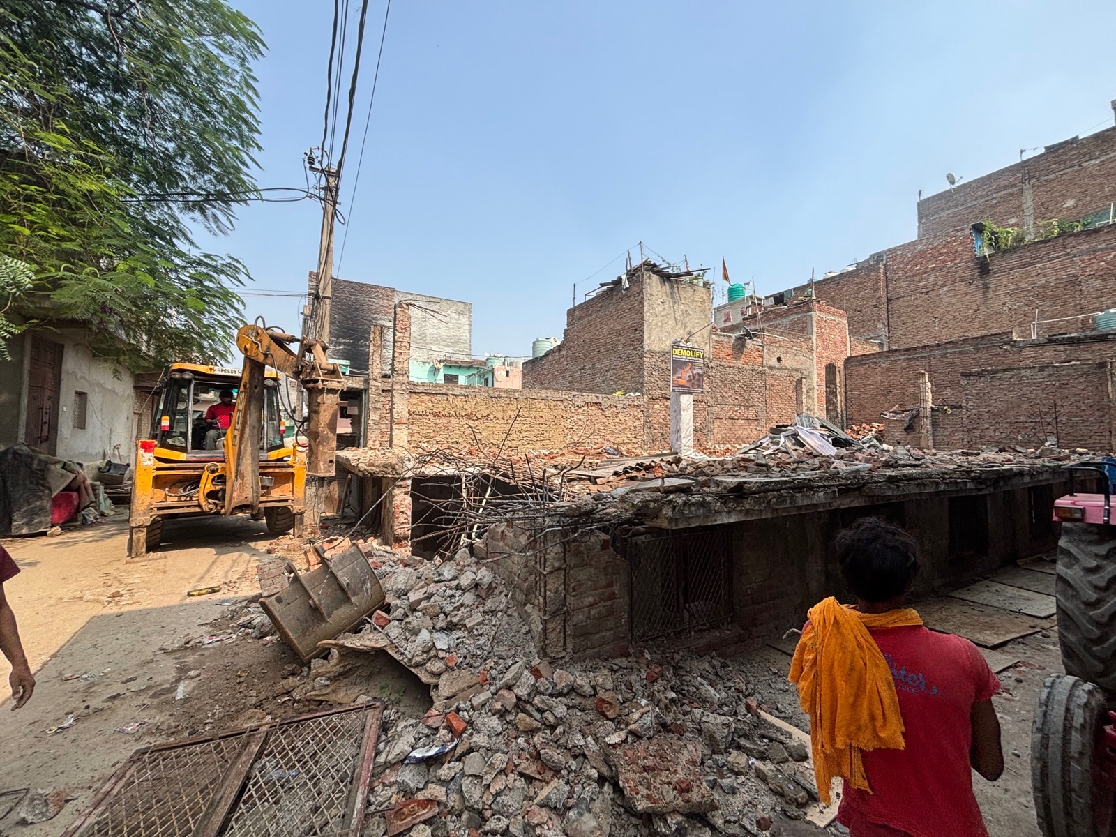JCB with a breaker demolishing a single-storey house in a narrow Delhi lane, with the Demolify site board on the wall behind
