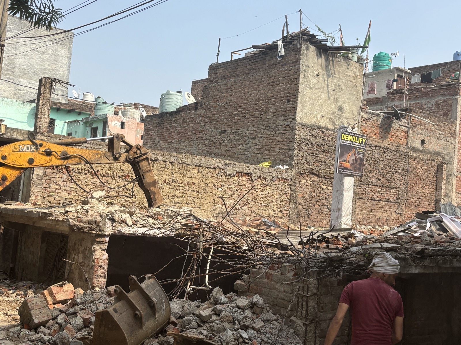 JCB breaker arm over a half-demolished house, with the Demolify site board on a pole and dense brick houses behind