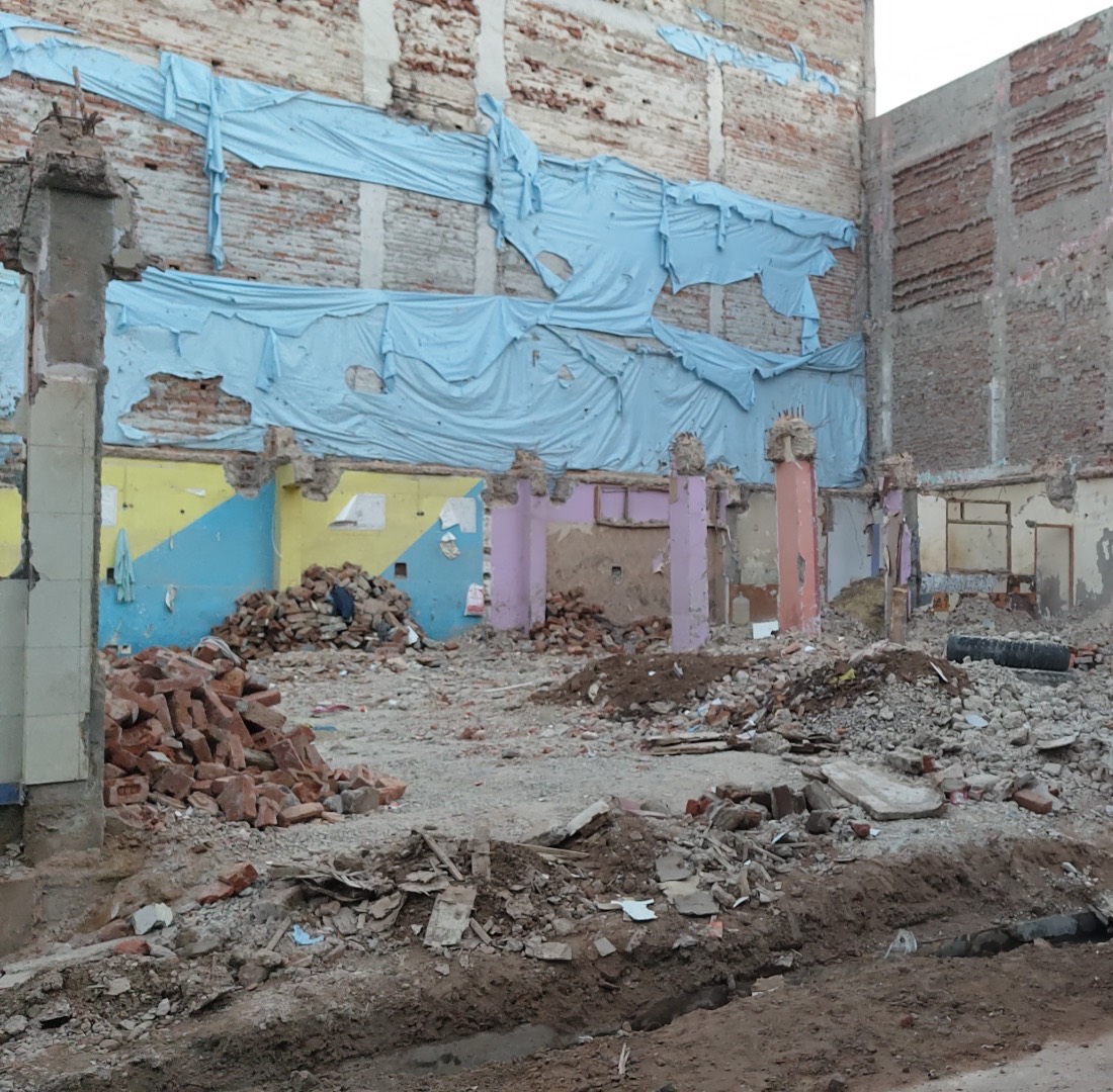 A cleared plot between old buildings, the neighbours' exposed walls covered with blue tarpaulin and good bricks stacked to one side