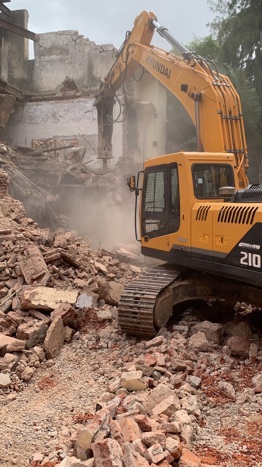 Hyundai excavator with a hydraulic breaker pulling down a brick house, dust rising from the rubble
